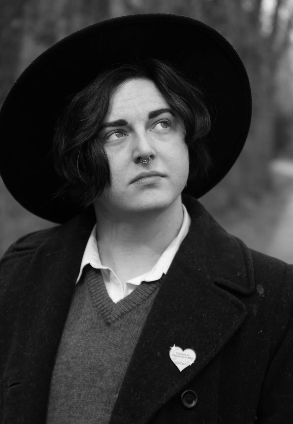 Author photo in black and white. The author is wearing a black felt hat, a peacoat, and a heart shaped pin from the Ripped Bodice